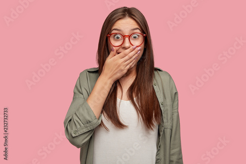 Fotografie, Obraz Isolated shot of happy European girl with minimal makeup, covers mouth as smiles or laughs at something funny, dressed in stylish shirt, isolated over pink background