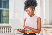 Way To Success. Young African American Woman With Afro Hairstyle Wearing Sleeveless Light Color Top, Standing In Vintage Office Building In New York, Looking Down, Working On Laptop Computer, Smiling