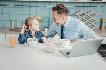 Wall Mural - Best father. Lovely attentive occupied father sitting by the table in the kitchen having breakfast and smiling to his daughter.