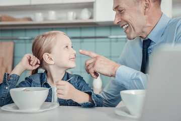 Wall Mural - Happy together. Pleasant smiling caring father sitting in the kitchen by the table looking at his daughter touching her nose.