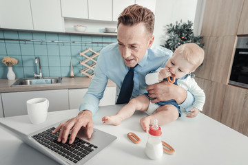 Wall Mural - Little helper. Pleasant busy concentrated man standing in the kitchen hugging a nursling who sitting on the table and using the laptop.