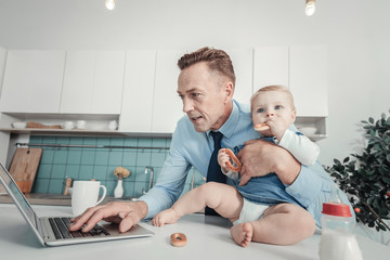 Wall Mural - Family business. Occupied caring handsome man standing in the kitchen near the table hugging a baby and working with the laptop.