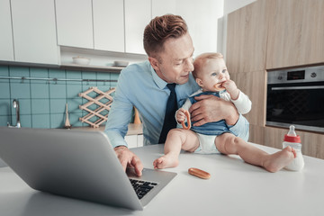 Wall Mural - Im with you. Pleasant lovely attentive father spending time in the kitchen looking at baby and using the laptop.