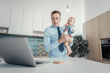 Wall Mural - Work will not wait. Serious concentrated busy man standing in the bright room holding a baby and looking at the laptop.