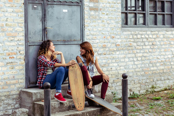 Two girl friends sitting at the stairs in front of metal door with skateboards and making fun