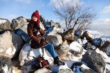 Wall Mural - Winter warm photo. Portrait of a beautiful young model in knitted hat  and warm clothes smilling and sitting on stone in the north pole, behind mountains covered in snow.  joyful cheerful mood.