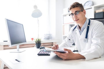 A young man in a white robe sitting at a table in the office. He holds a pen in his hand and looks straight. The portrait of the doctor.