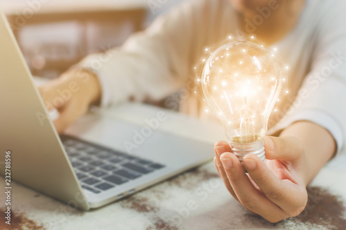 Fotomural Woman hand holding light bulb and using laptop on wooden desk