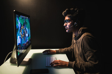 Gamer or streamer in earphones with microphone sits at home in dark room and plays with friends on networks in video games. A young man sits in front of a monitor.