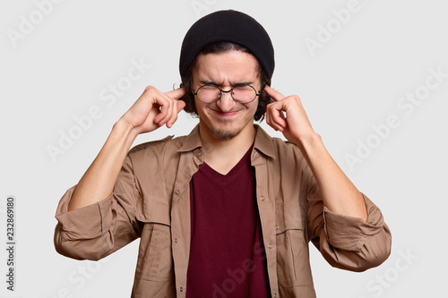 Fotografía Stressful teenager wears black har and beige shirt, pluggs ears, ignores loud sound coming from noise neighbours, stands against white background