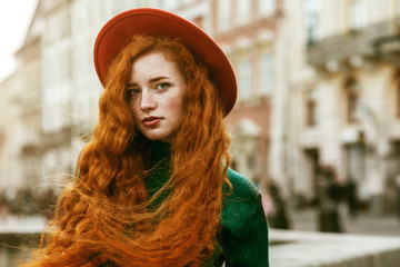 Wall Mural - Close up portrait of young beautiful fashionable redhead woman with freckles, very long curly hair, wearing green turtleneck, orange hat, posing in street of european city. Copy, emty space for text