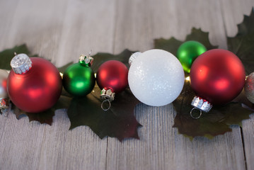 Group of 3 red ornaments, 2 green ornaments, one white ornament on green leaves on a light wood background