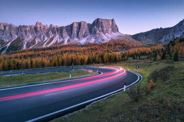 Wall Mural - Blurred car headlights on winding road in mountains at sunset in autumn. Spectacular landscape with asphalt road, light trails, forest, rocks and purple sky at night in fall. Car driving on roadway