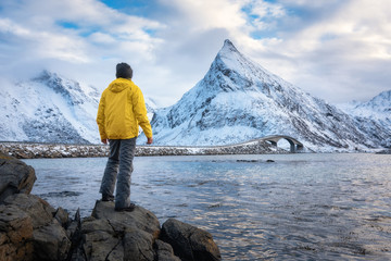 Wall Mural - sporty man in yellow jacket standing in the stone on seacoast against snowy mountains and cloudy sky