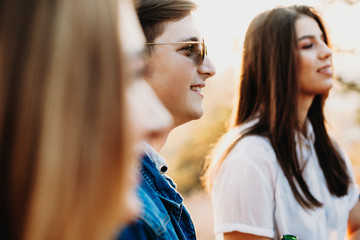 Side view of attractive guy in stylish glasses smiling and looking away while standing in countryside near female friends. Smiling man near friends in nature