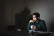 © ChayTee - Attractive young asian man drinking coffee sitting on desk table looking at laptop computer in dark late night working feeling serious thinking and determinated at home office in work hard concept.