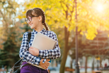 Happy 12 years old teenager girl in eyeglasses with two braids holding workbooks looking aside in the autumn park after school. Copy space.