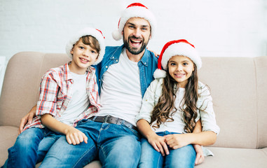 Wall Mural - Handsome father with him son and daughter. Happy young family in red santa hats spending time together at home while winter holidays