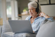 © goodluz - Senior woman at home on laptop with headphones