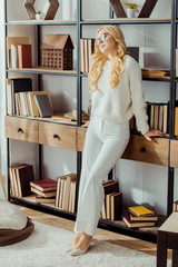 Wall Mural - attractive woman in glasses standing near rack with books in living room