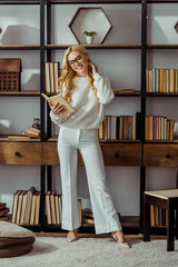 Wall Mural - attractive woman in glasses standing near rack and reading book in living room