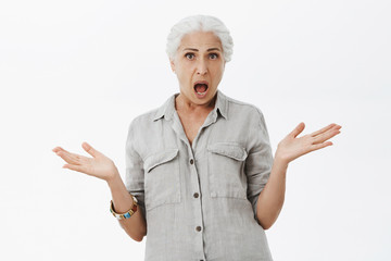 Studio shot of confused and displeased shocked senior mother with white hair shrugging raising palms aside questioned arguing with kid being clueless why he become so naughty over gray wall