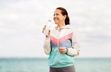 fitness, sport and healthy lifestyle concept - woman drinking water after exercising at seaside