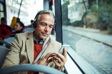 mature tired businessman with heaphones and smartphone travelling by bus in city.