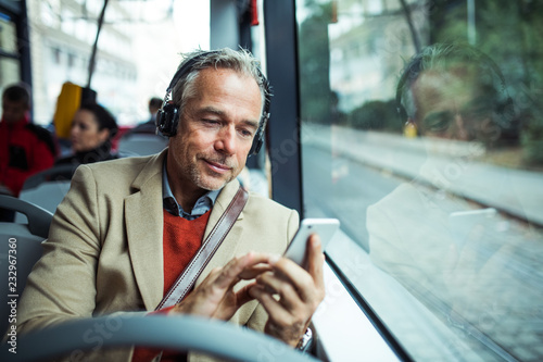 Fotomural Mature tired businessman with heaphones and smartphone travelling by bus in city