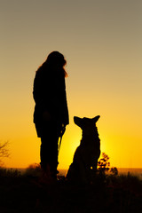 silhouette woman with a dog looking at each othe rin the field at sunset, pet sitting near girl's leg on nature,