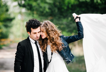 Cute couple embraces. Portrait of stylish young man in suit with tie hugs beautiful girl holding white dress in air. Concept of successful relationship and happy moments. Fashion wedding photography