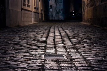 Empty alley paved with wet cobblestone at night illuminated by street lamps with a sewer cap in focus