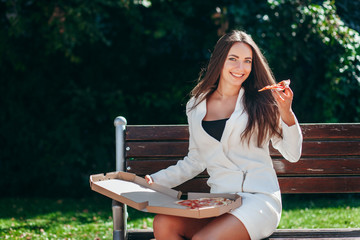 Smiling business girl in white dress eating pizza in street 