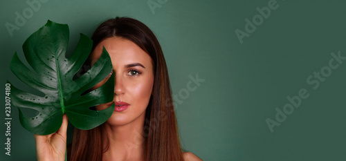 Fotografía Young woman with natural make up and tropical monstera leaf in hand over green background with copy space