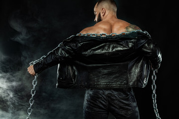 Wall Mural - Men fashion. Close-up portrait of a brutal bearded man topless in a leather jacket with chains. Athlete bodybuilder on black background.