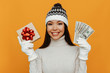 Christmas presents. Woman portrait. Accessories. Asian girl in a white polo neck, cap and gloves is holding a gift box and money and smiling, on an orange background