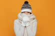 Woman portrait. Accessories. Warmness. Asian girl in a white scarf, cap and gloves is showing she is cold, on an orange background