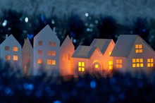 Handmade Small White Cardboard Houses With Illuminated Windows On Dark Blue Bokeh Foreground And Background. Winter Decoration. Shallow Depth Of Focus.