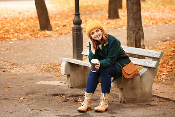 Sticker - Beautiful young woman with coffee resting on wooden bench in autumn park