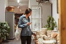 Young Woman With Mobile Phone Choosing Crockery Arranged On Table In Boutique