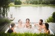 © Maskot - Cheerful male and female friends enjoying drinks in hot tub against lake during weekend getaway