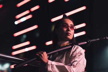 low angle view of attractive asian girl in white kimono holding japanese sword on street with neon light in evening, city of future concept