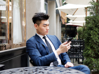 Portrait of a handsome businessman in blue suit using mobile phone at a restaurant outdoor, close up upper body view. Attractive male hipster Chinese model.