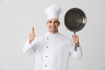 Wall Mural - Emotional young man chef indoors isolated over white wall background holding frying pan.