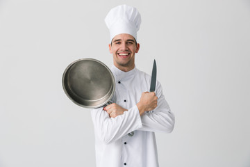 Wall Mural - Emotional young man chef indoors isolated over white wall background holding frying pan.