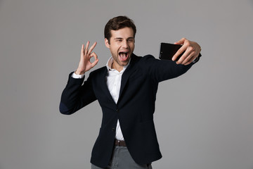 Canvas Print - Image of joyous businessman 30s in formal suit showing ok sign while taking selfie photo on cell phone, isolated over gray background