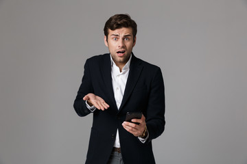 Poster - Image of stressful man 30s in business suit using black smartphone, isolated over gray background