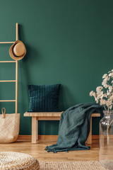 Pillow and blanket on bench in green apartment interior with plant, pouf and hat on ladder. Real photo