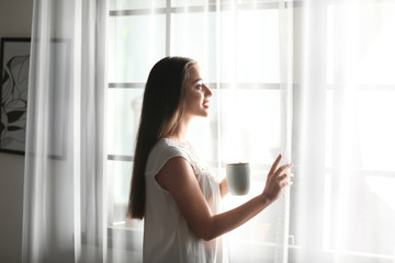 Sticker - Young woman standing near window with curtains at home