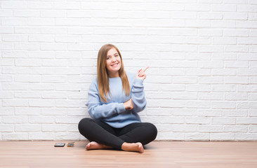 Sticker - Young adult woman sitting on the floor in autumn over white brick wall with a big smile on face, pointing with hand and finger to the side looking at the camera.